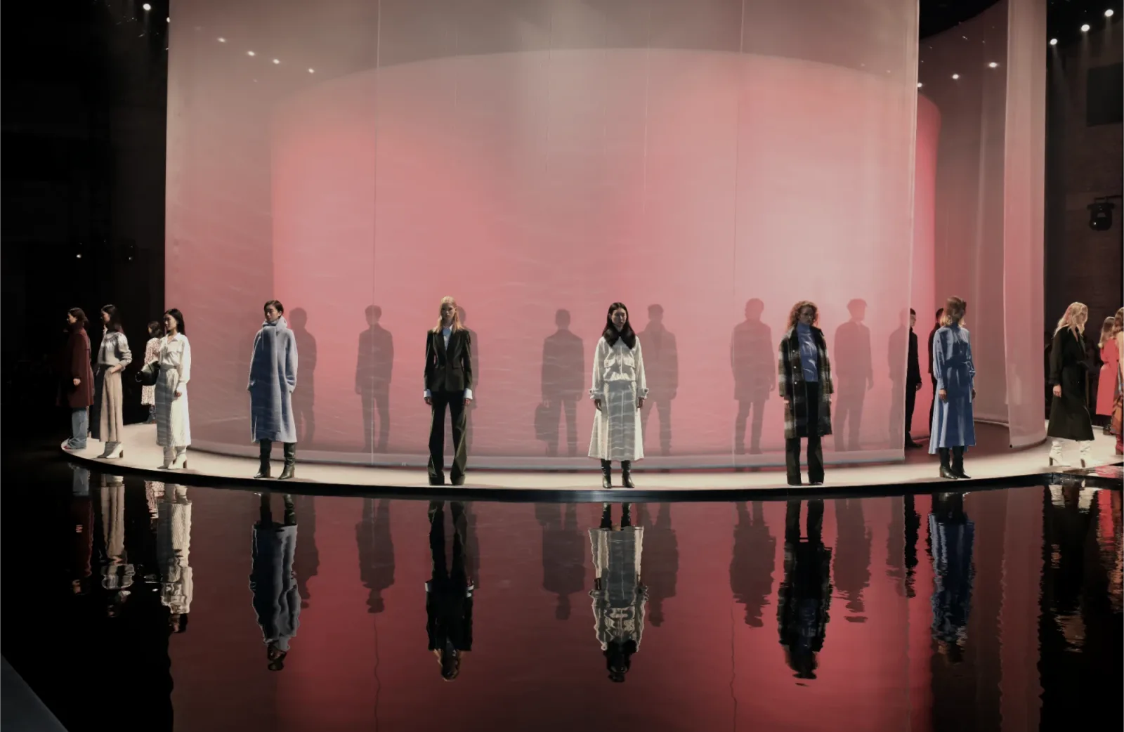 Models standing on a circular runway before a translucent pink cylinder, mirrored in a reflective water surface at a Massimo Dutti fashion show.