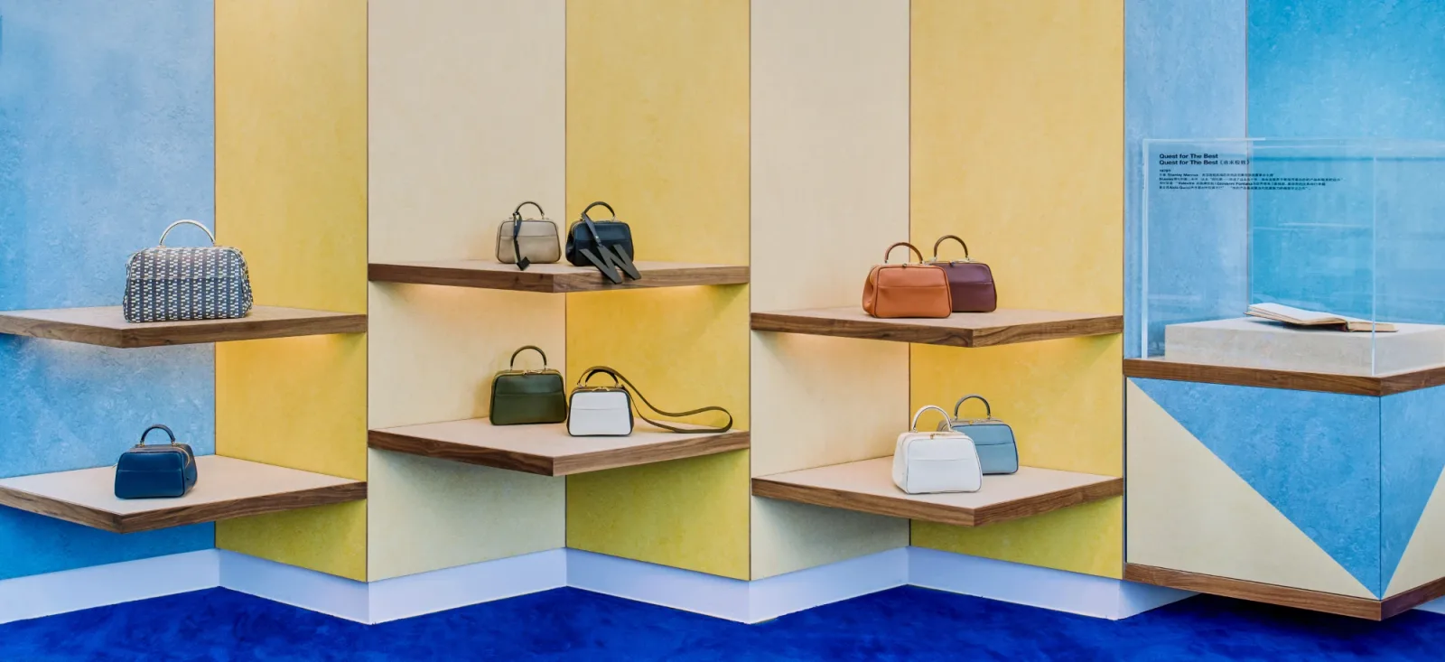 Valextra handbags arranged on angled timber shelves against blue and yellow walls above a cobalt carpet.