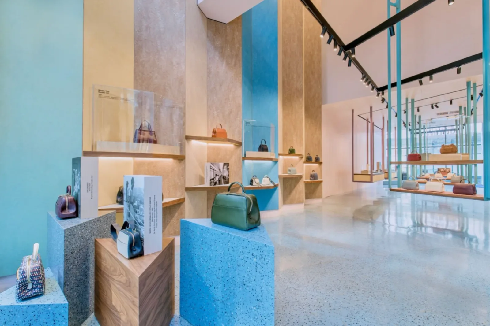 Valextra store interior with leather bags displayed on terrazzo plinths and timber shelves against blue and sand-coloured walls.